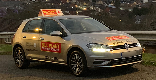 silver driving school car parked on a road with illuminated lights and a rooftop sign featuring the letter L indicating learner driving lessons in a residential area at dusk providing essential driving skills for 11 students