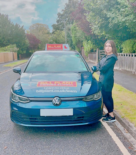 woman standing next to blue Volkswagen driving school car with L plate in residential area focused on learning to drive 4 driving lessons