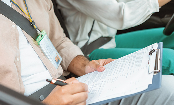person holding clipboard and writing in car with passenger wearing seatbelt focused on paperwork for driving test